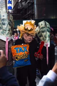 A papier-mâché Donald Trump wearing a crown, and surrounded by dollar bills, and holding a packet of Cheetos that reads "Tax Cheato President".