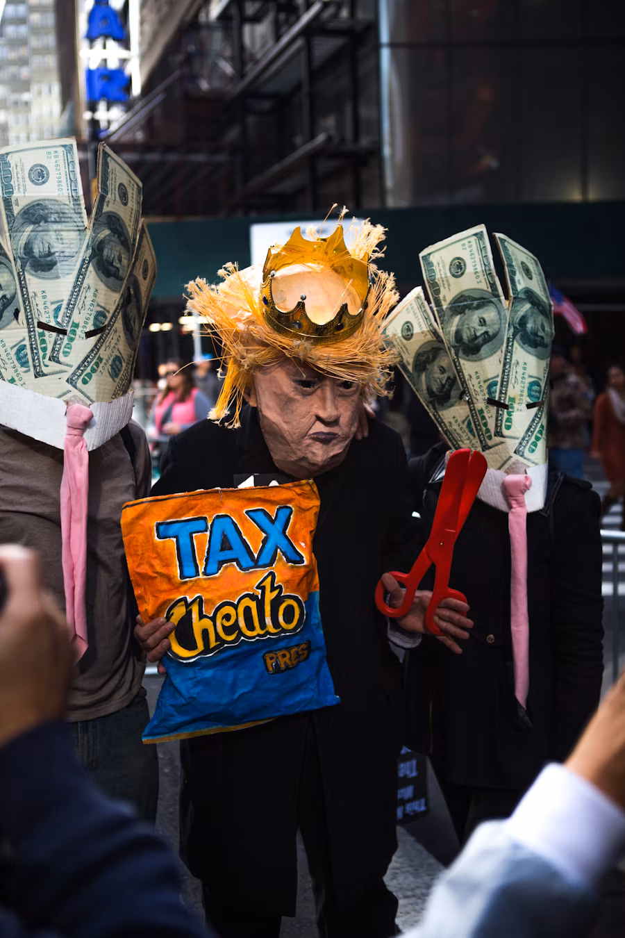 A papier-mâché Donald Trump wearing a crown, and surrounded by dollar bills, and holding a packet of Cheetos that reads "Tax Cheato President".